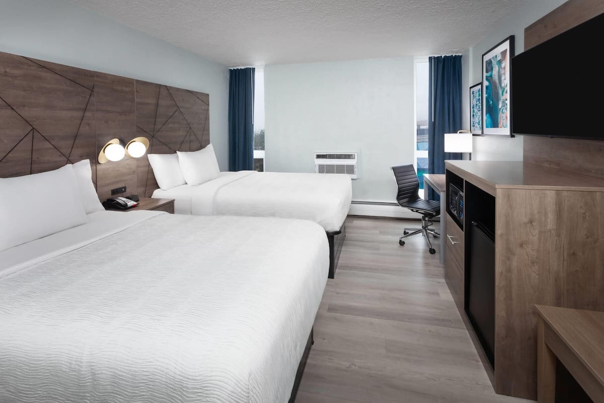 A hotel room with two beds, a desk, a chair, a TV, and an air conditioning unit.