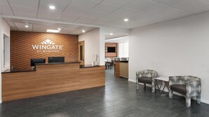 Uma área de recepção com balcão de madeira, um espaço de estar com duas cadeiras e uma mesa, e uma cozinha ao fundo.