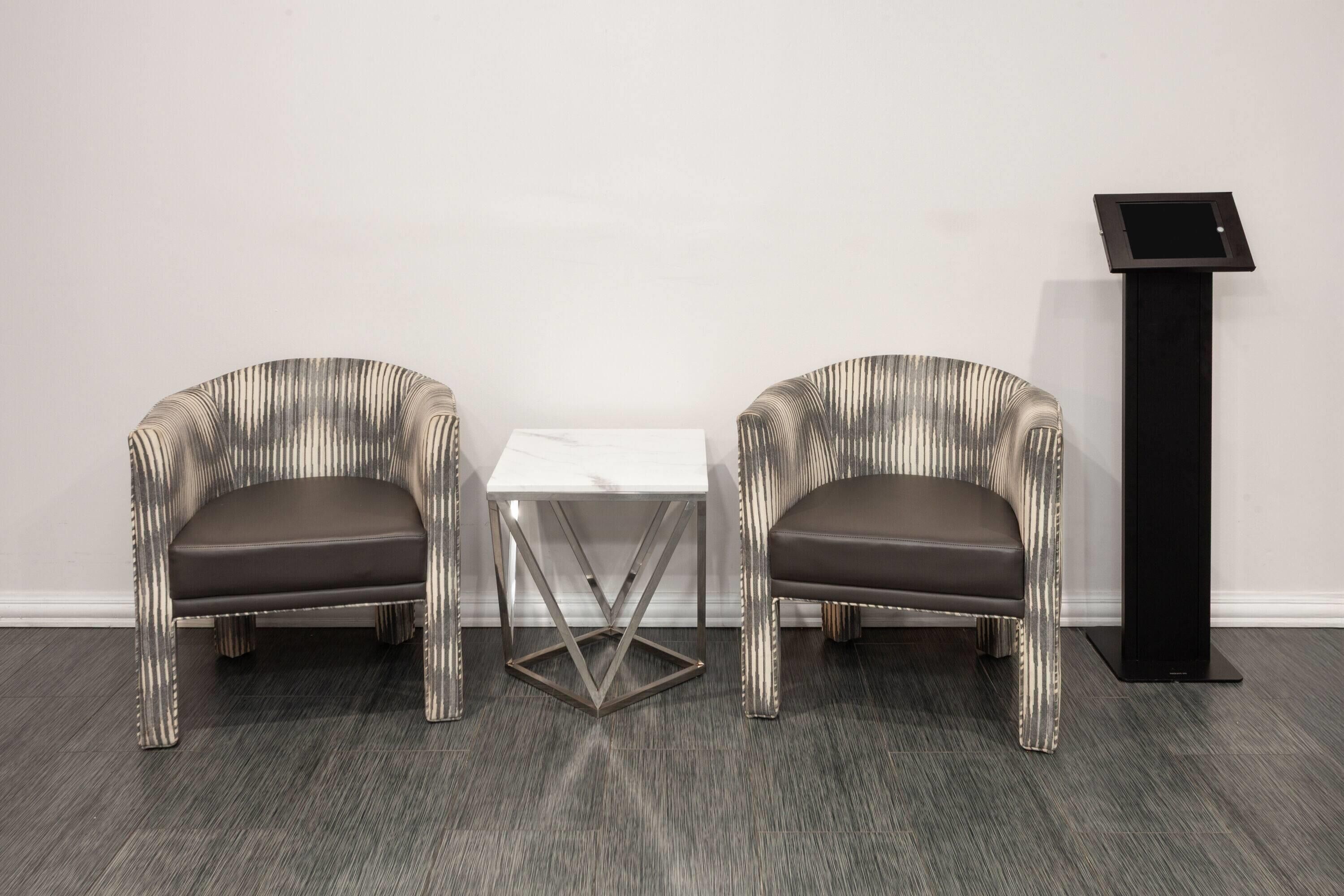 Twee gestoffeerde stoelen met metalen poten, een klein wit tafeltje met een metalen voet en een zwarte verticale standaard met een scherm.