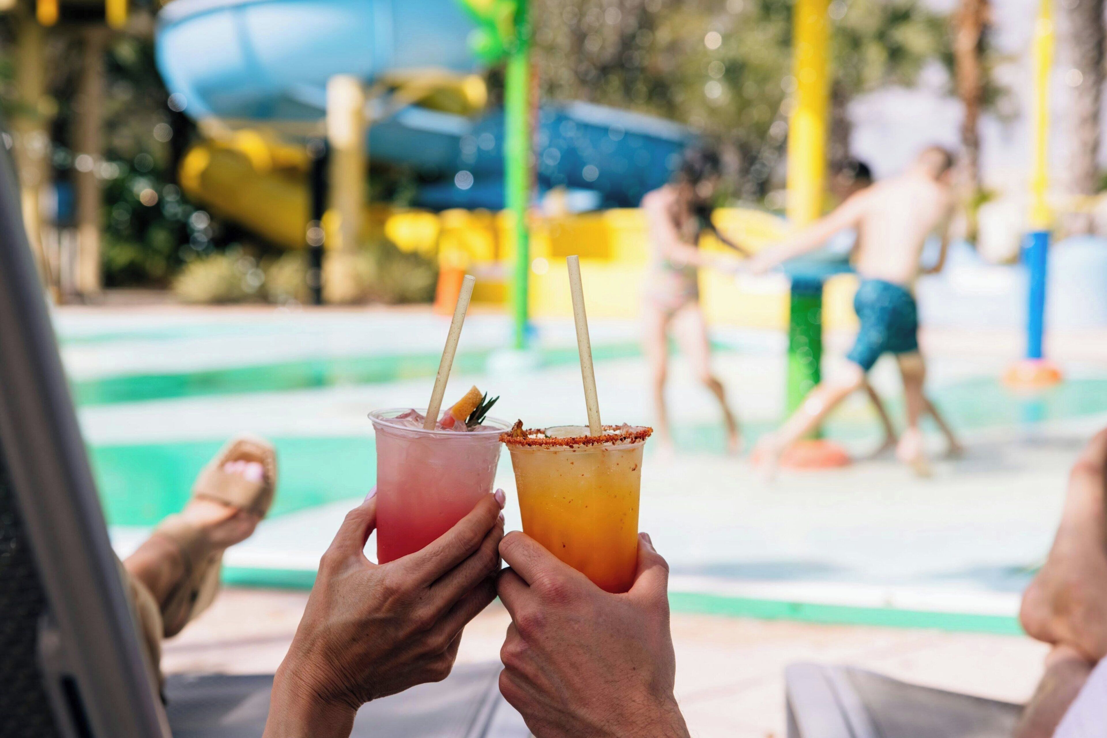 Two people holding drinks at a poolside area with a water slide in the background.