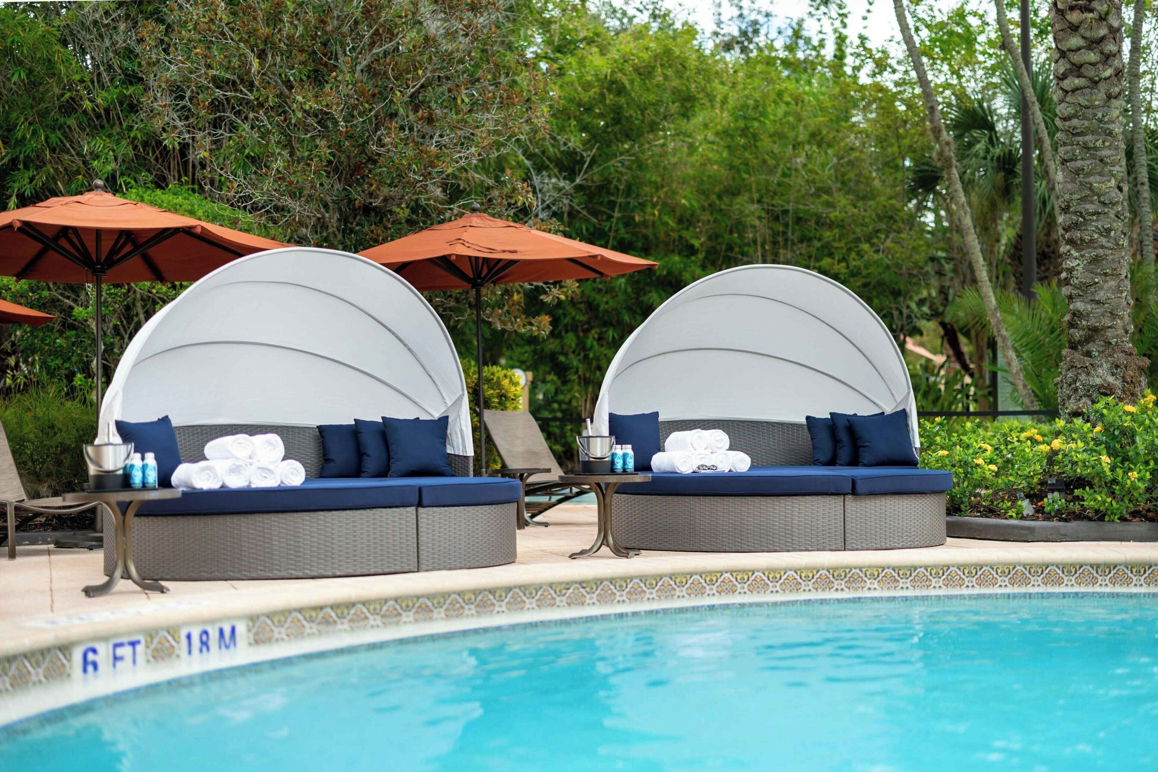 Deux chaises longues avec parasols, installées au bord d’une piscine, entourées de verdure.