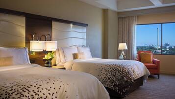 A hotel room with two beds, a nightstand with lamps, a chair, and a large window with a city view.