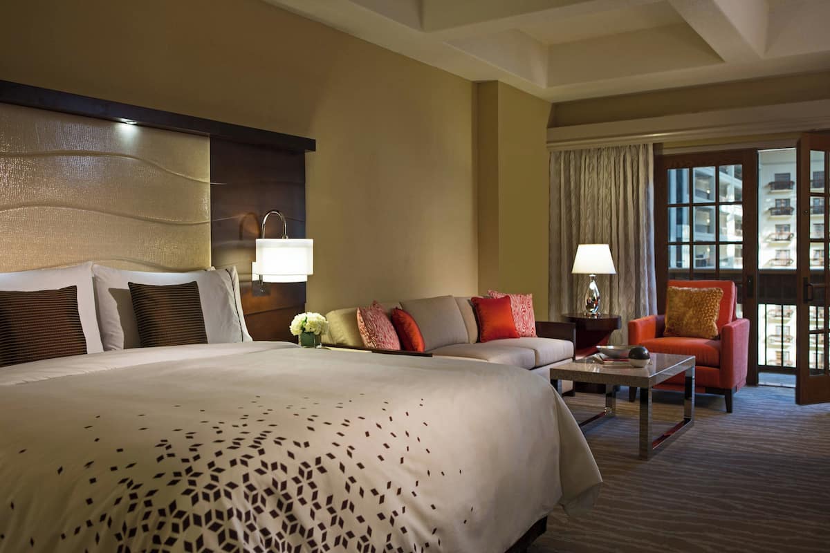 Hotelkamer met een groot bed, een bank, een salontafel en een stoel. Er is een raam met gordijnen en uitzicht op gebouwen.