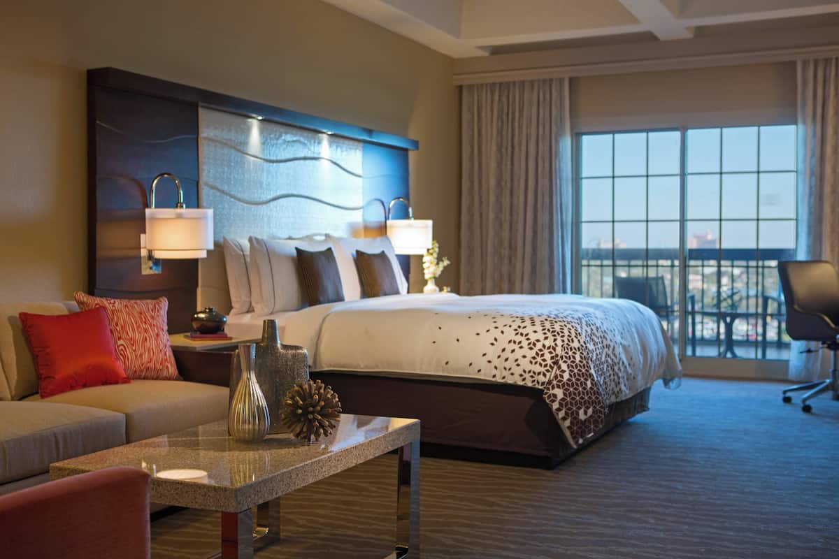 Een hotelkamer met een groot bed, twee bedlampen, een nachtkastje, een salontafel en uitzicht op het landschap door een groot raam.