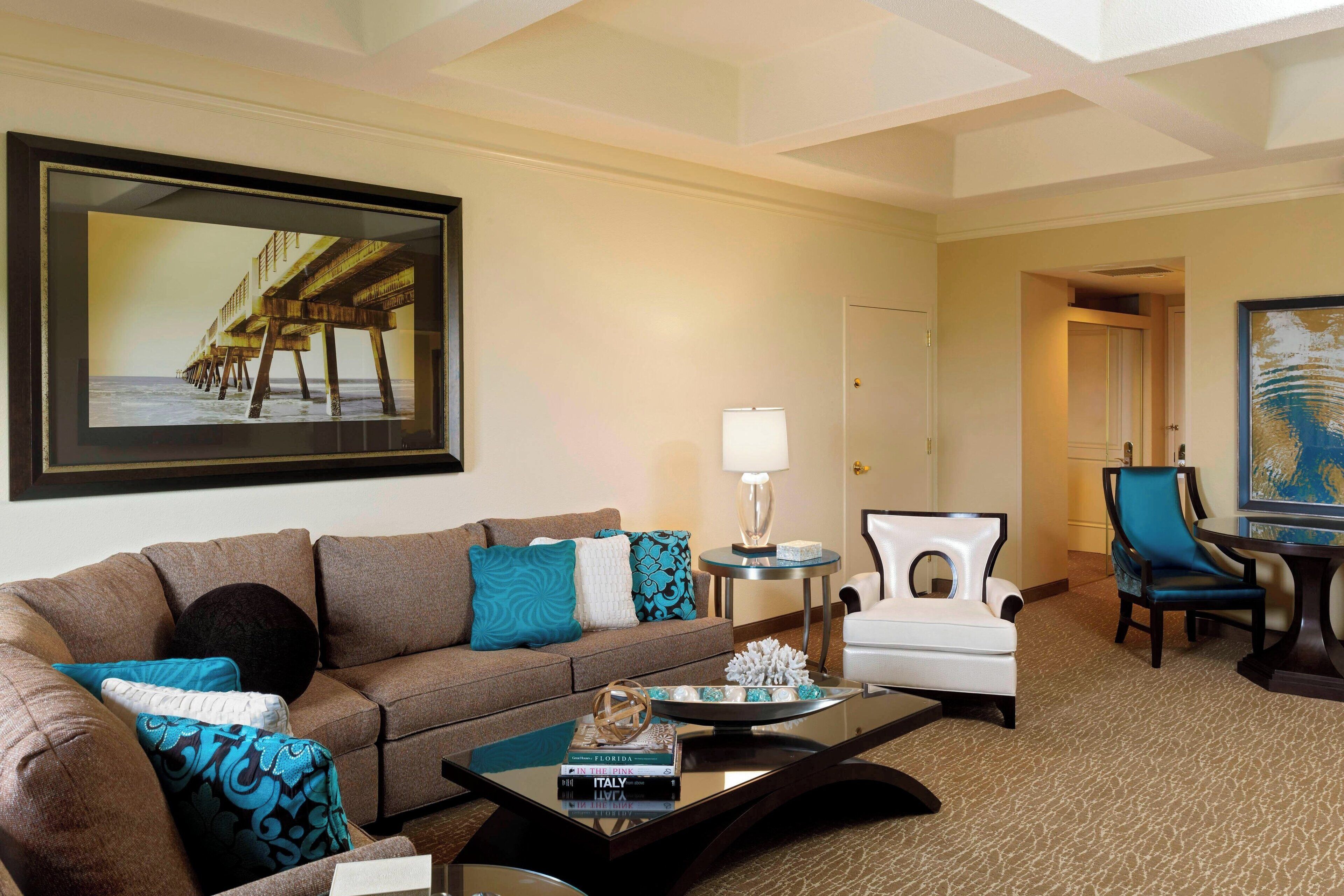 A living room with a brown sofa, teal pillows, a coffee table with books, a lamp, and a framed picture of a pier.