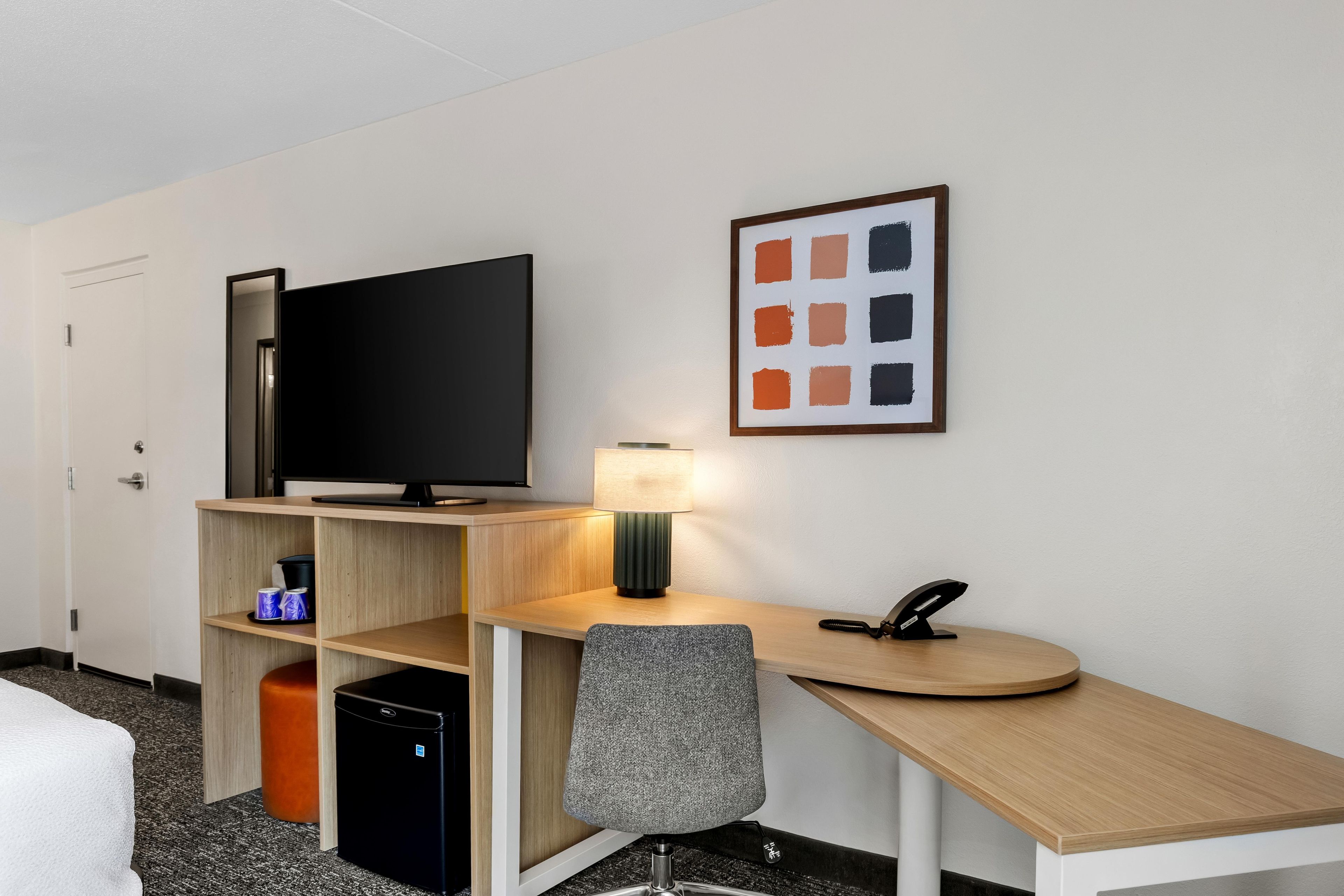 Habitación de hotel con televisor de pantalla plana, escritorio con lámpara, silla, teléfono y un cuadro en la pared.