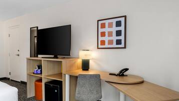 Habitación de hotel con televisor de pantalla plana, escritorio con lámpara, silla, teléfono y un cuadro en la pared.