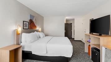 A hotel room with a bed, bedside lamps, a flat-screen TV, a small refrigerator, and a door leading to another room.