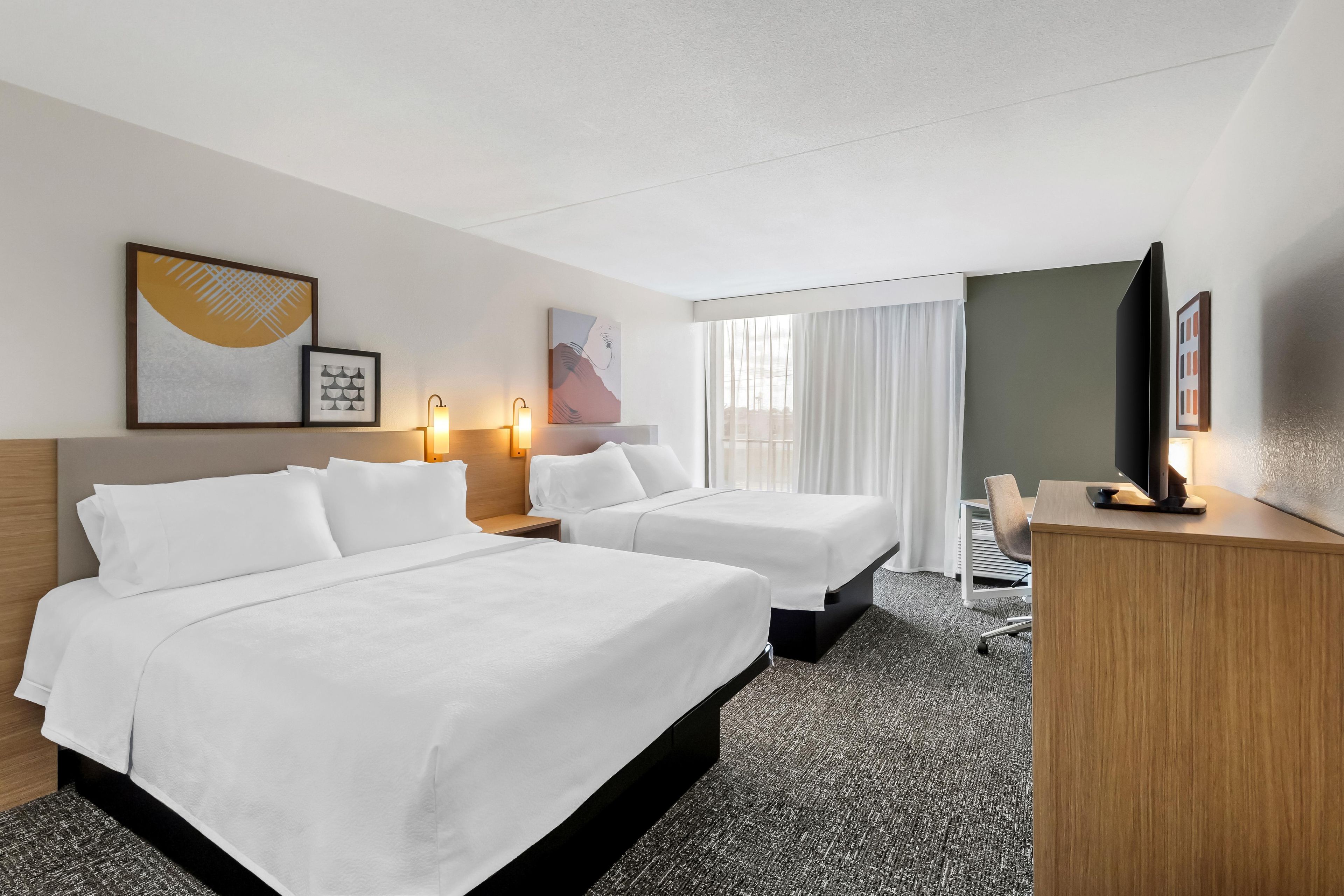 Habitación de hotel con dos camas, un escritorio, un televisor y obras de arte en las paredes.