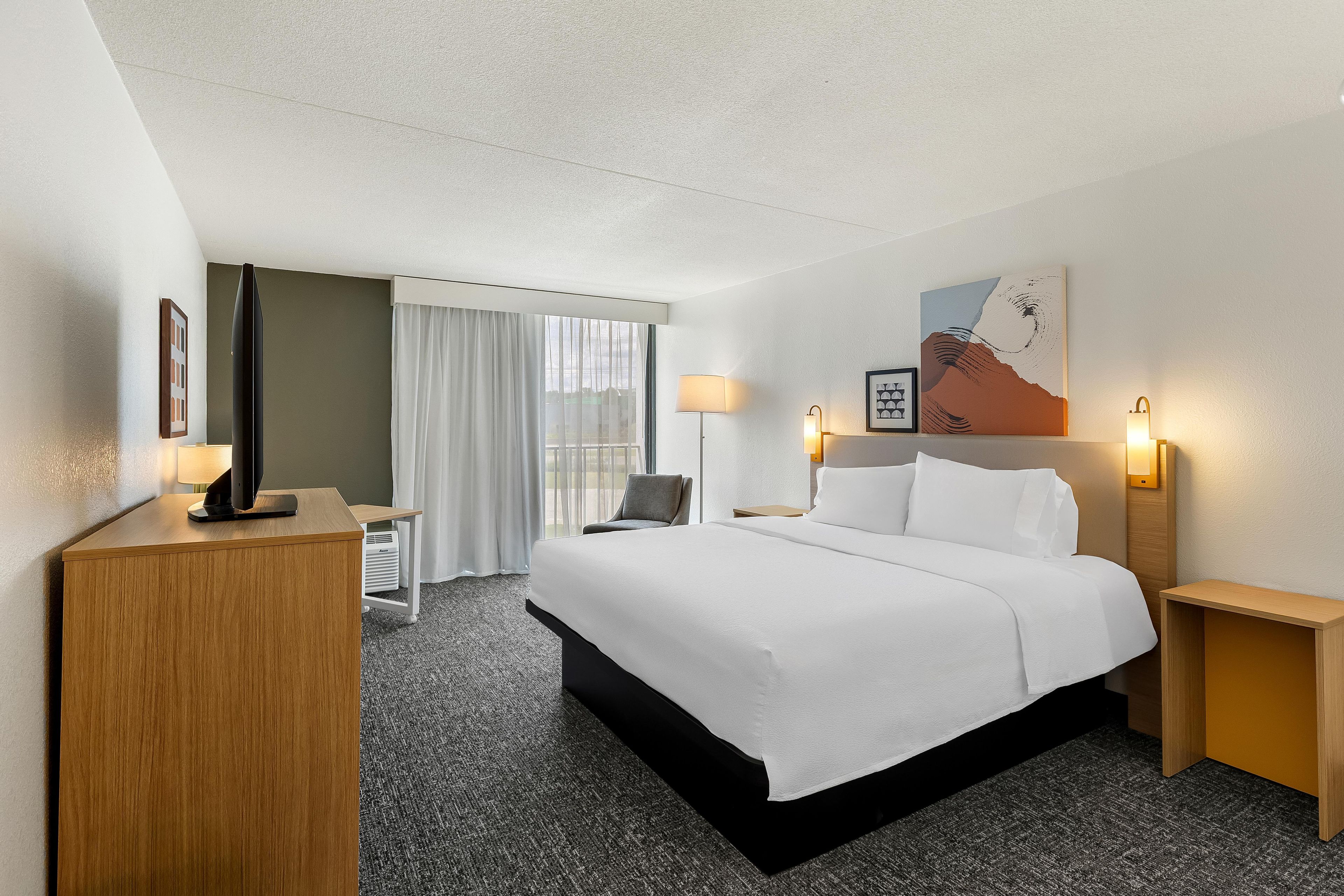 A hotel room with a bed, bedside tables, a desk, a chair, and a window with curtains.