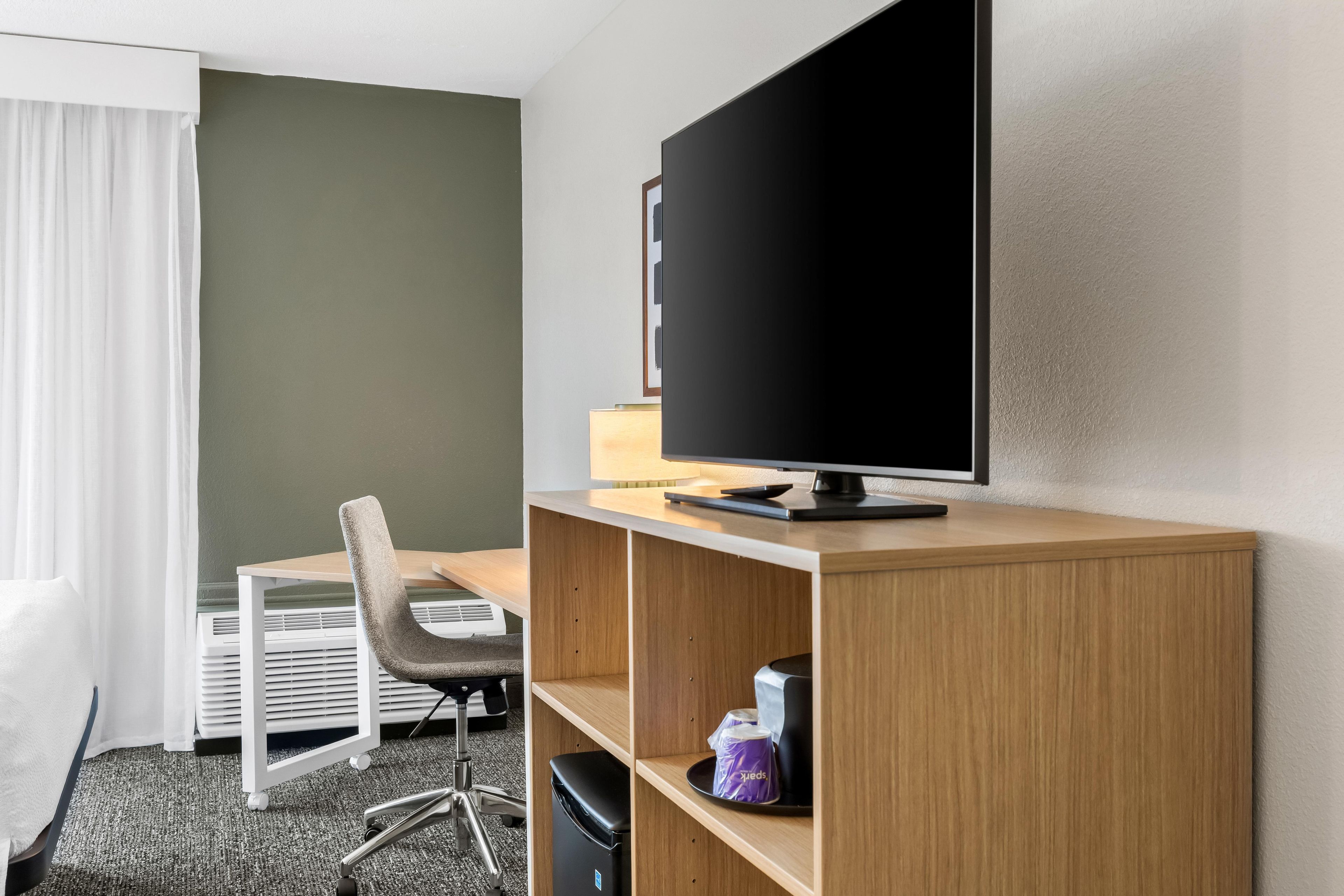 Una habitación de hotel con escritorio, silla, televisión y un estante con objetos.
