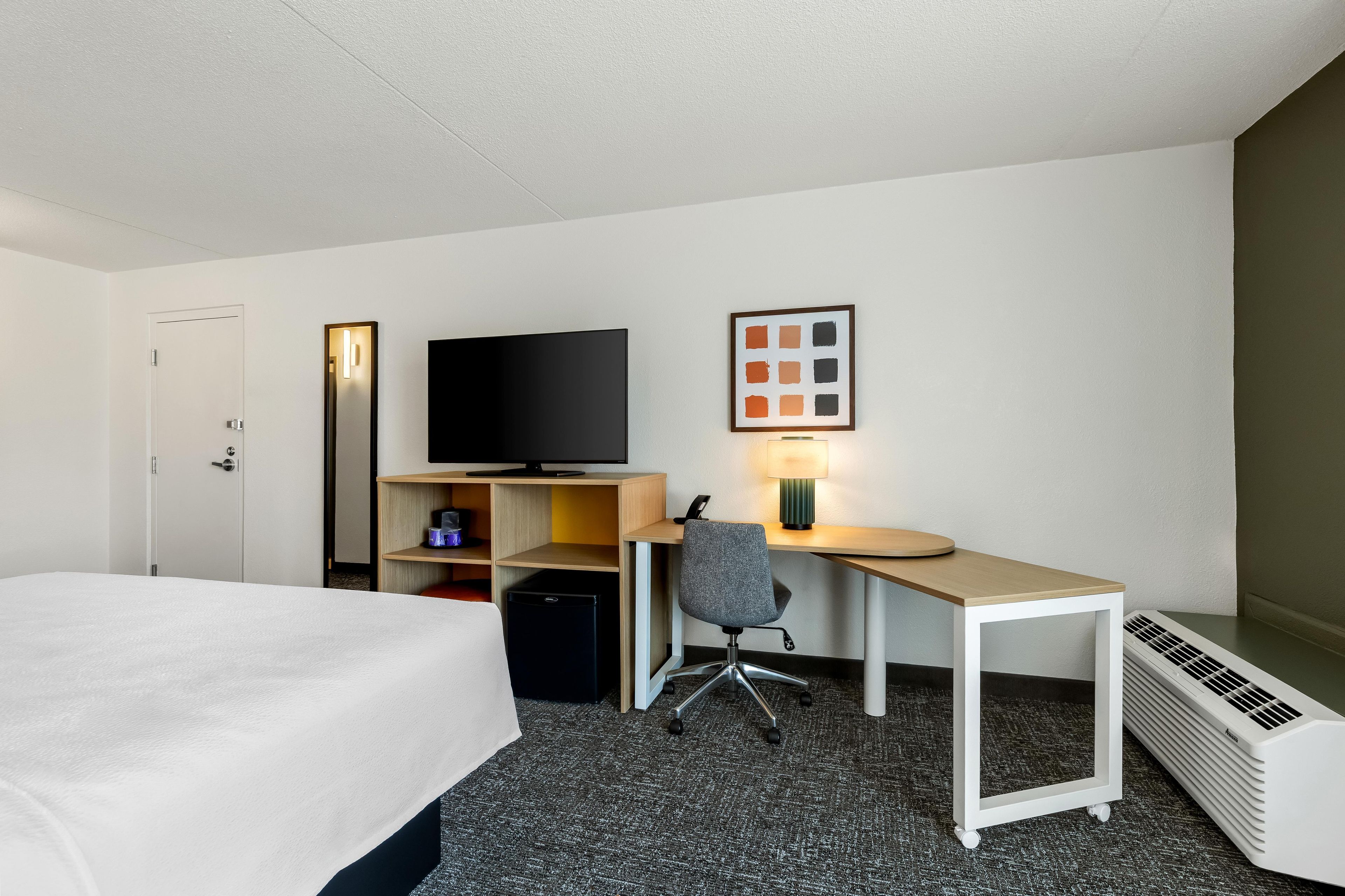 Habitación de hotel con cama, escritorio con silla, televisión, calefactor pequeño y un cuadro en la pared.