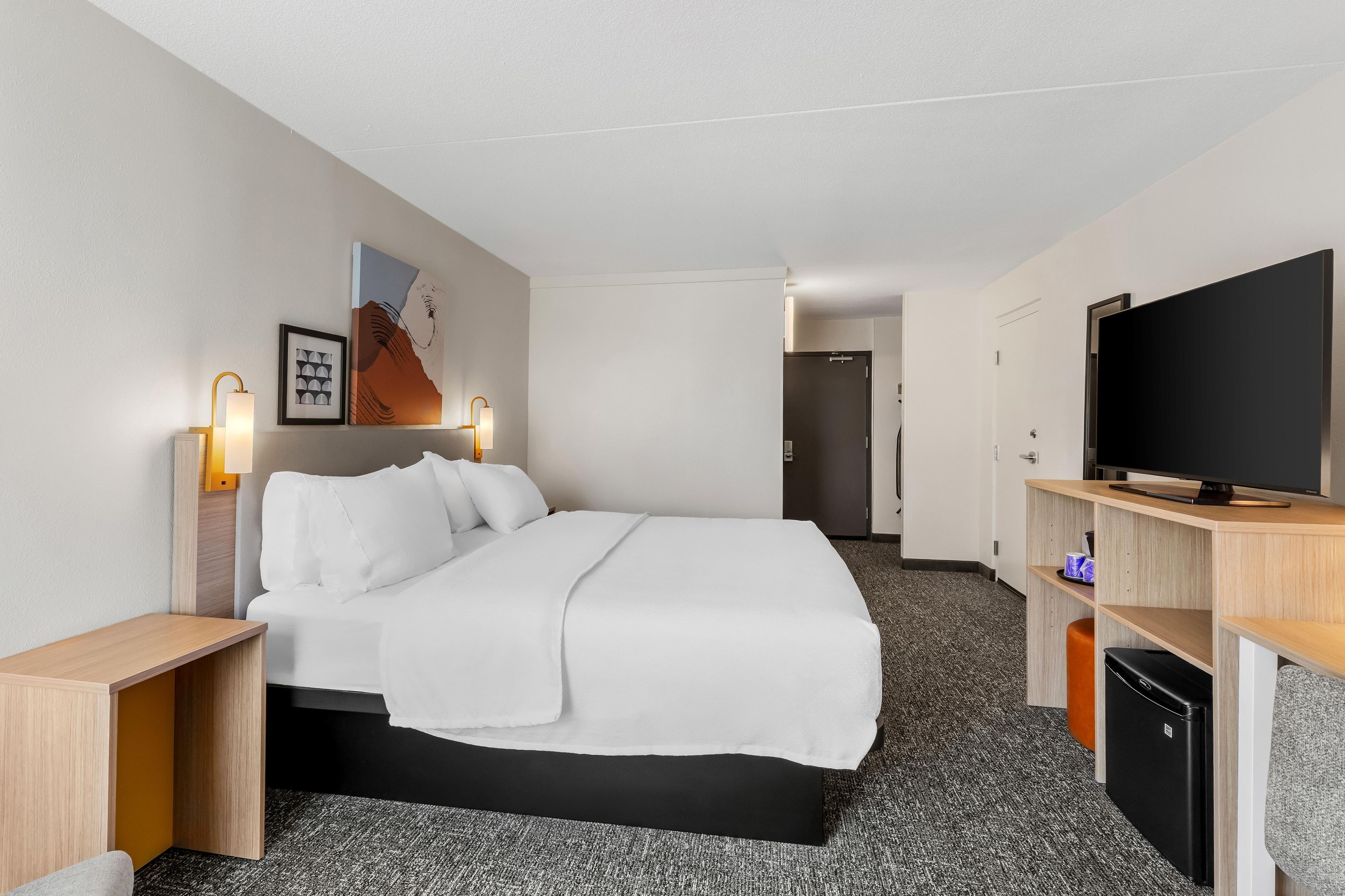 A hotel room with a bed, bedside lamps, a flat-screen TV, a small refrigerator, and a door leading to another room.
