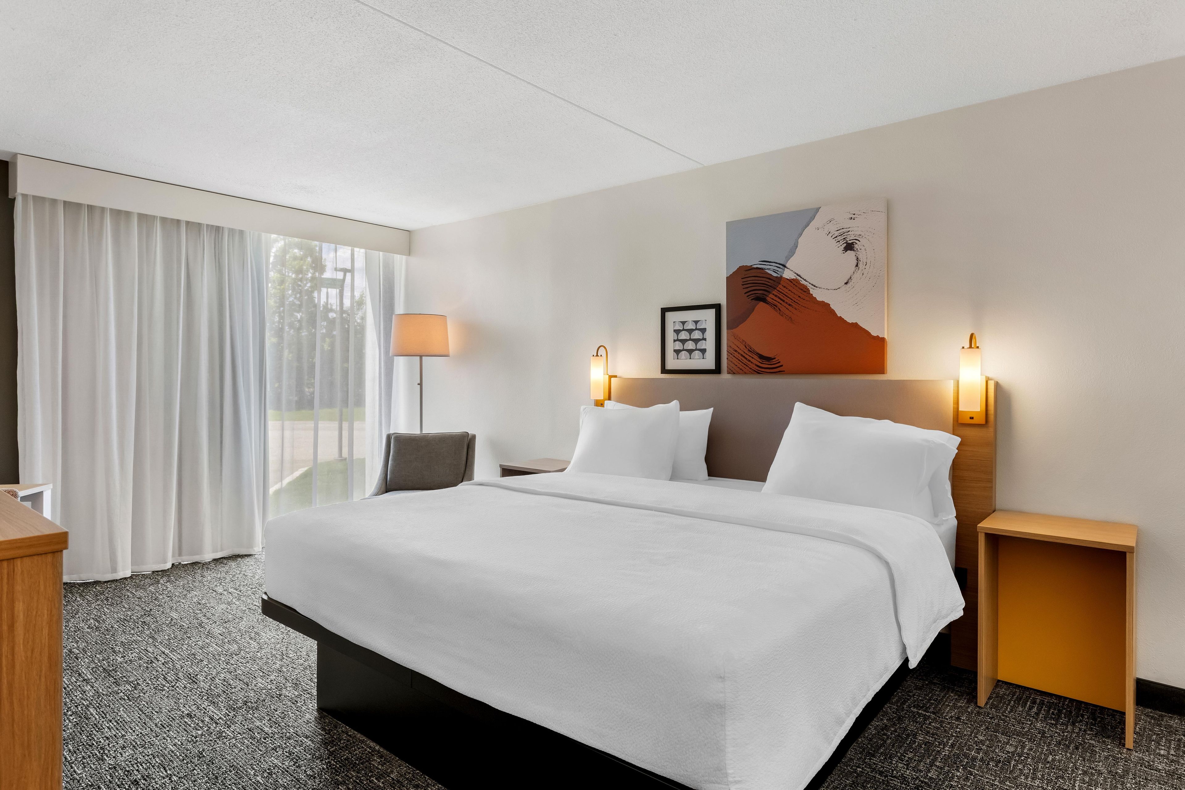 Habitación de hotel con una cama grande, un escritorio, una silla, una ventana con cortinas y una obra de arte en la pared.