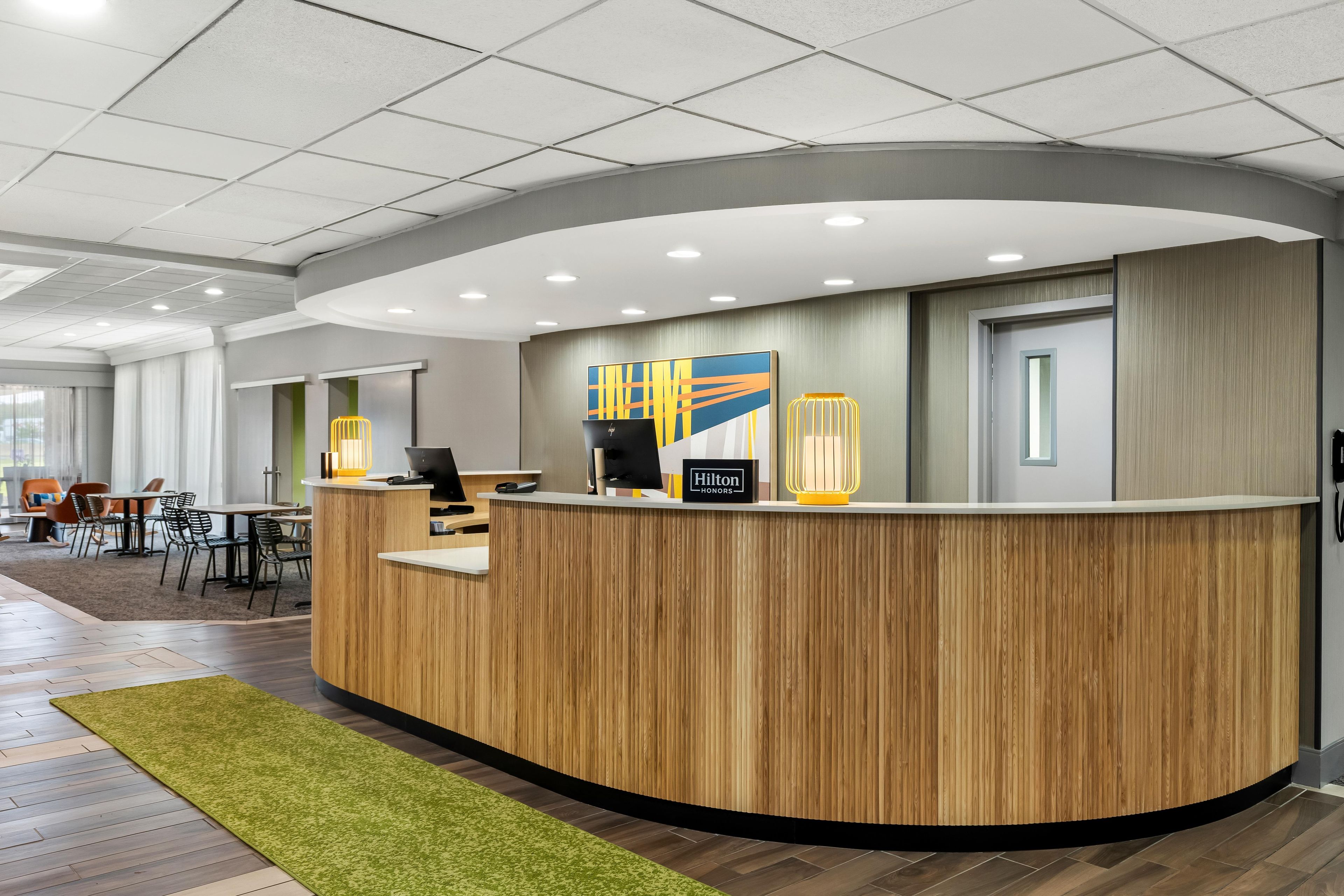 A hotel reception desk with a wooden finish, a large painting above it, and a seating area with tables and chairs in the background.
