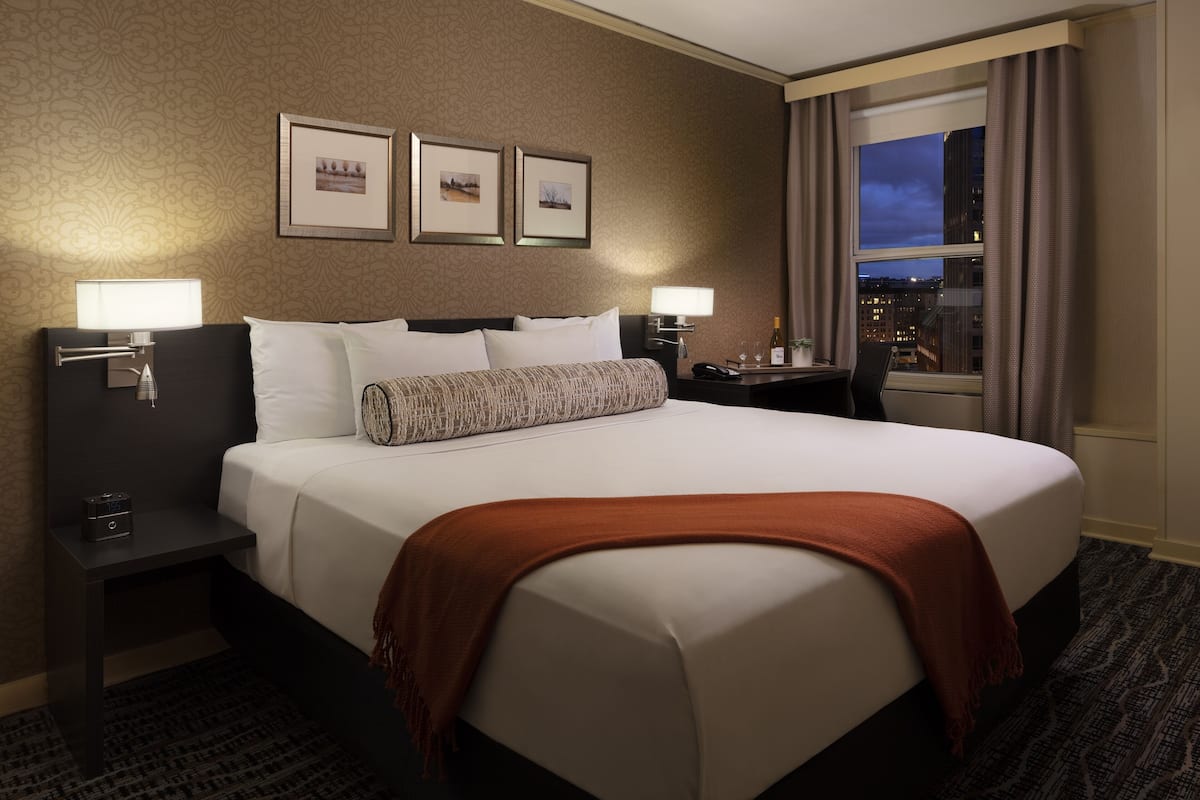 A hotel room with a large bed, two bedside tables, a nightstand, a window with a city view, and framed pictures on the wall.