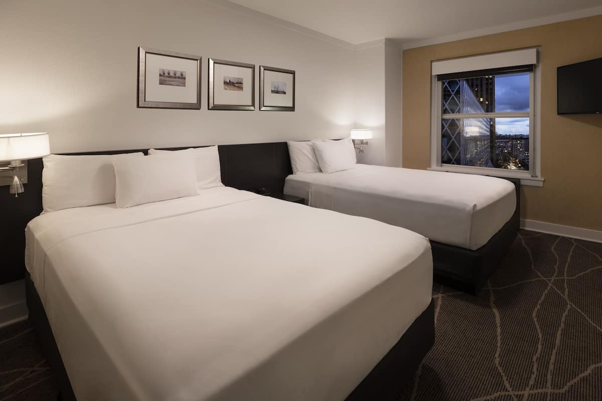 A hotel room with two beds, a large window with a city view, framed pictures on the wall, and a flat-screen TV mounted on the wall.