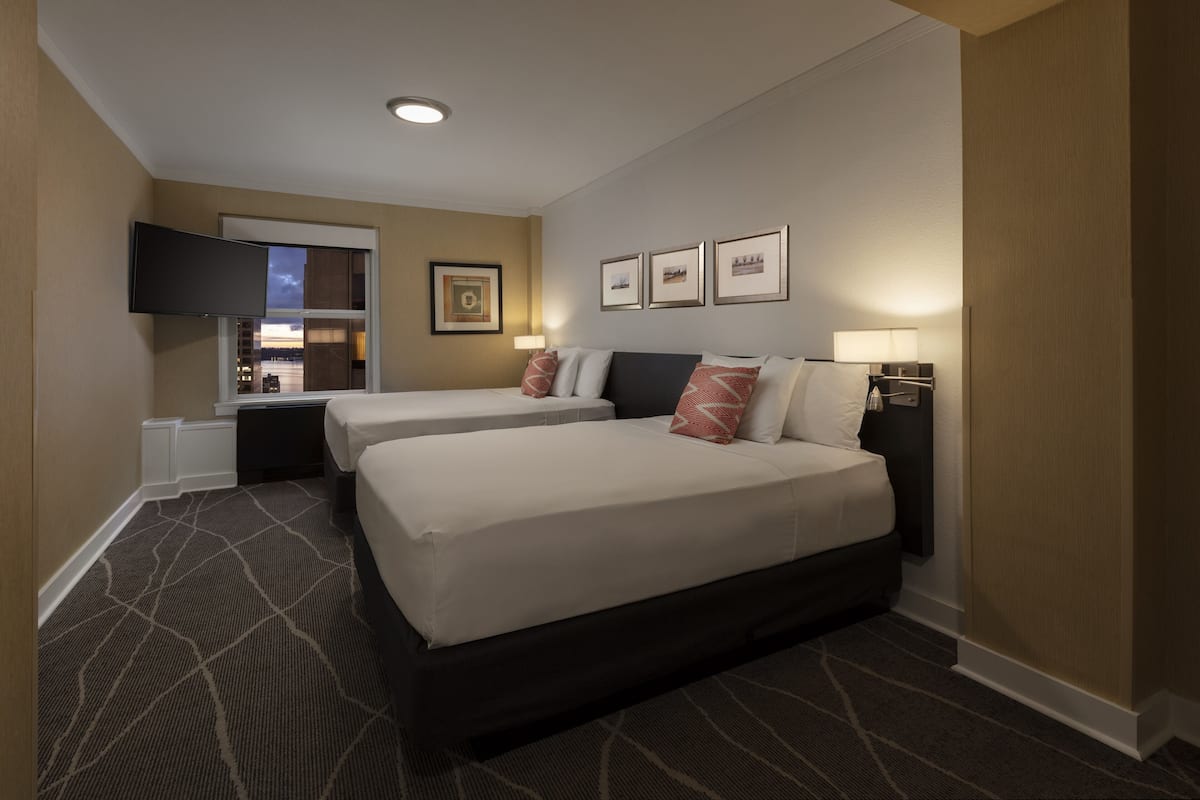 A hotel room with two beds, a flat-screen TV mounted on the wall, and a window with a view of the cityscape.
