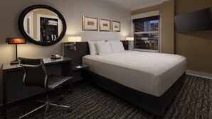 A hotel room with a bed, desk, chair, desk lamp, bedside lamps, a round mirror, framed pictures, a flat-screen TV, and a window with a city view.