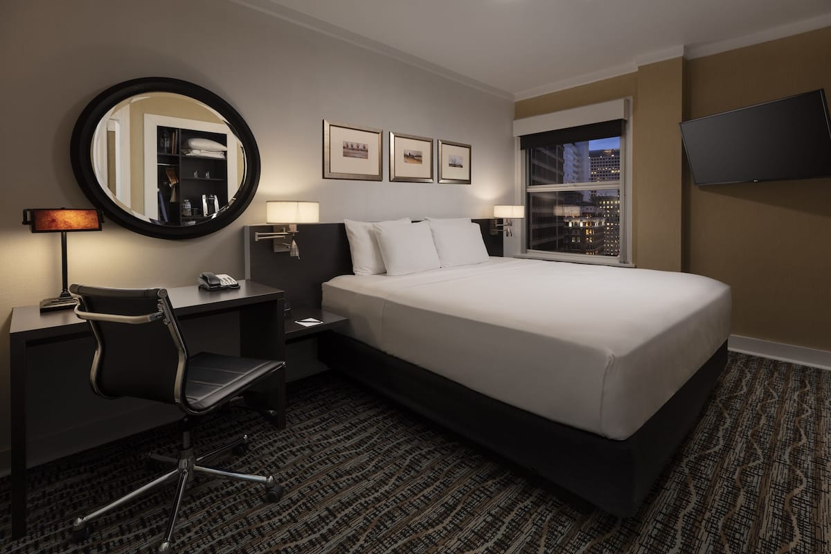 A hotel room with a bed, desk, chair, desk lamp, bedside lamps, a round mirror, framed pictures, a flat-screen TV, and a window with a city view.