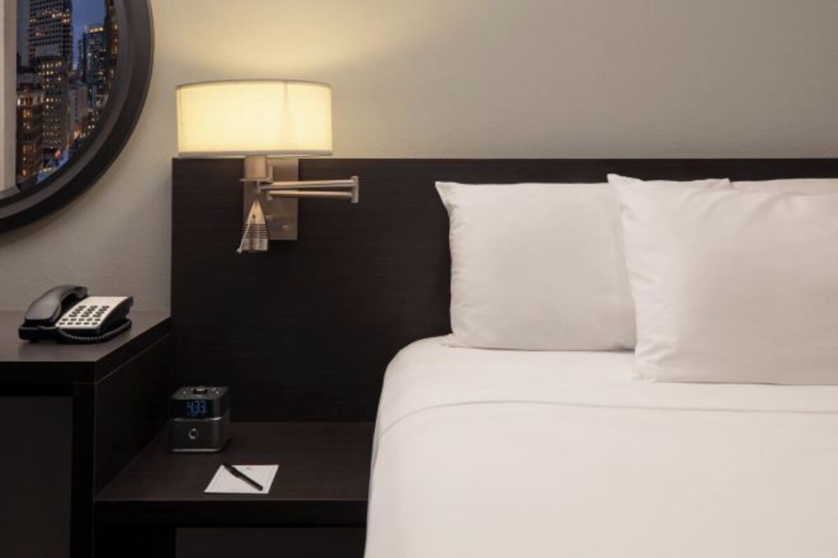 A hotel room with a bed, a nightstand with a lamp, a phone, and a digital clock.