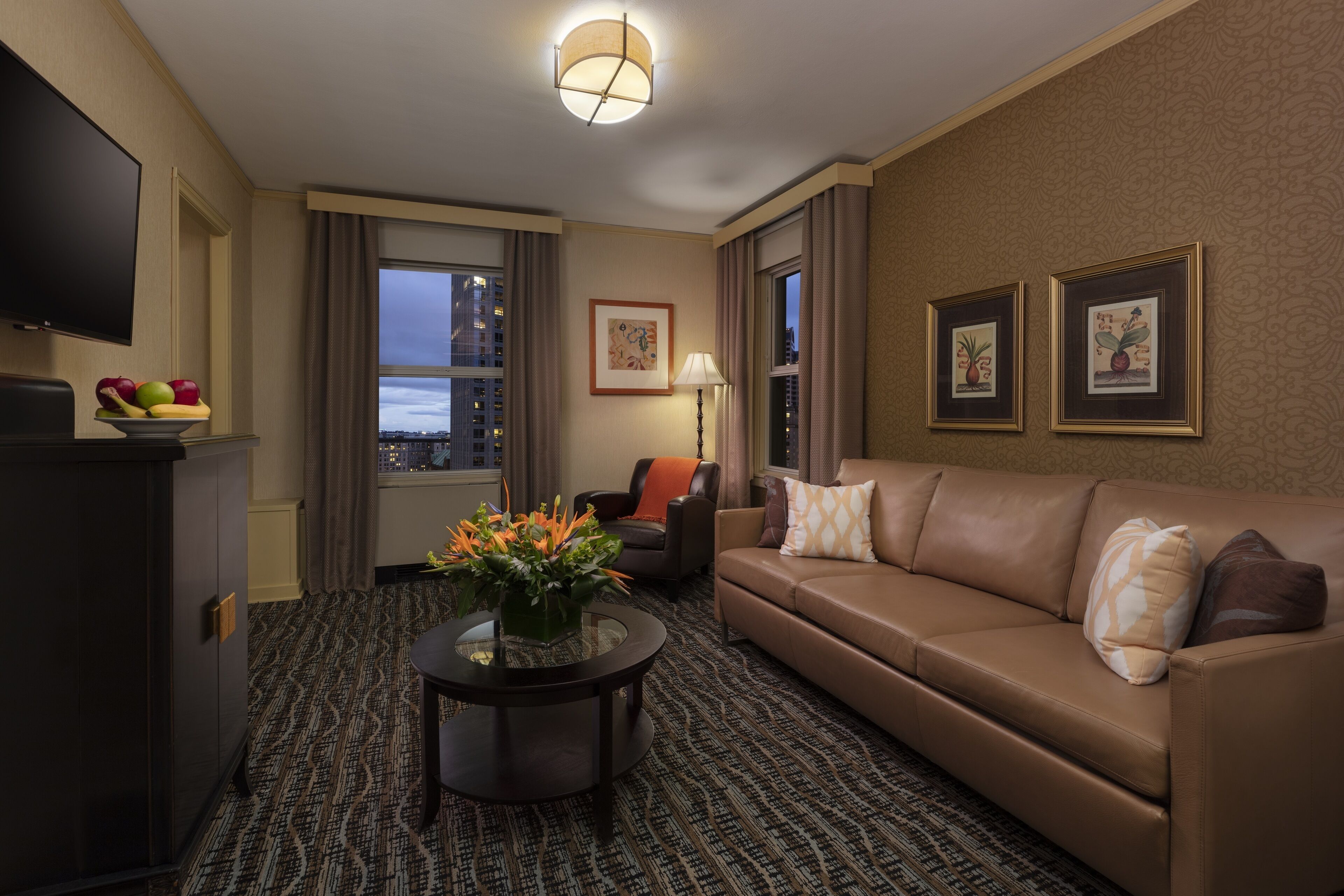 A hotel room with a leather sofa, a flat-screen TV, a coffee table with fruit, a city view, and framed artwork on the wall.