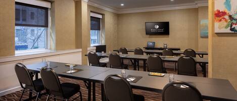A conference room with multiple tables and chairs, a flat-screen TV mounted on the wall, and a window with a view of buildings outside.