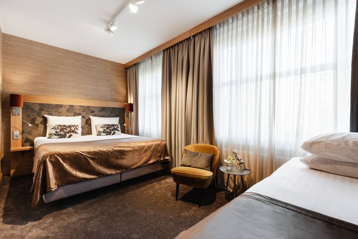A hotel room with a double bed, a single bed, a small armchair, a nightstand, and a lamp.