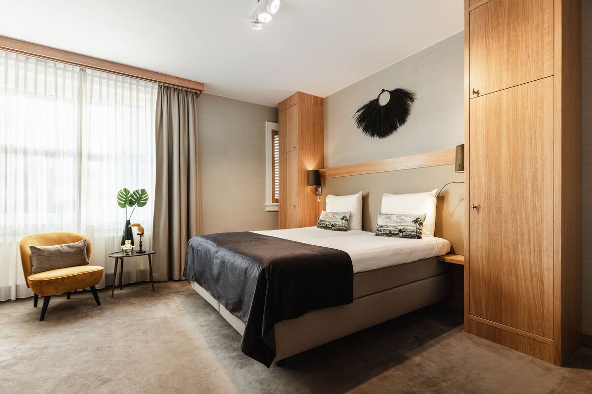 A modern hotel room with a large bed, a wooden wardrobe, a small table with a plant, and a comfortable armchair.