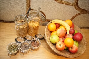 A wooden table with jars of granola, a bowl of apples and oranges, and a banana.