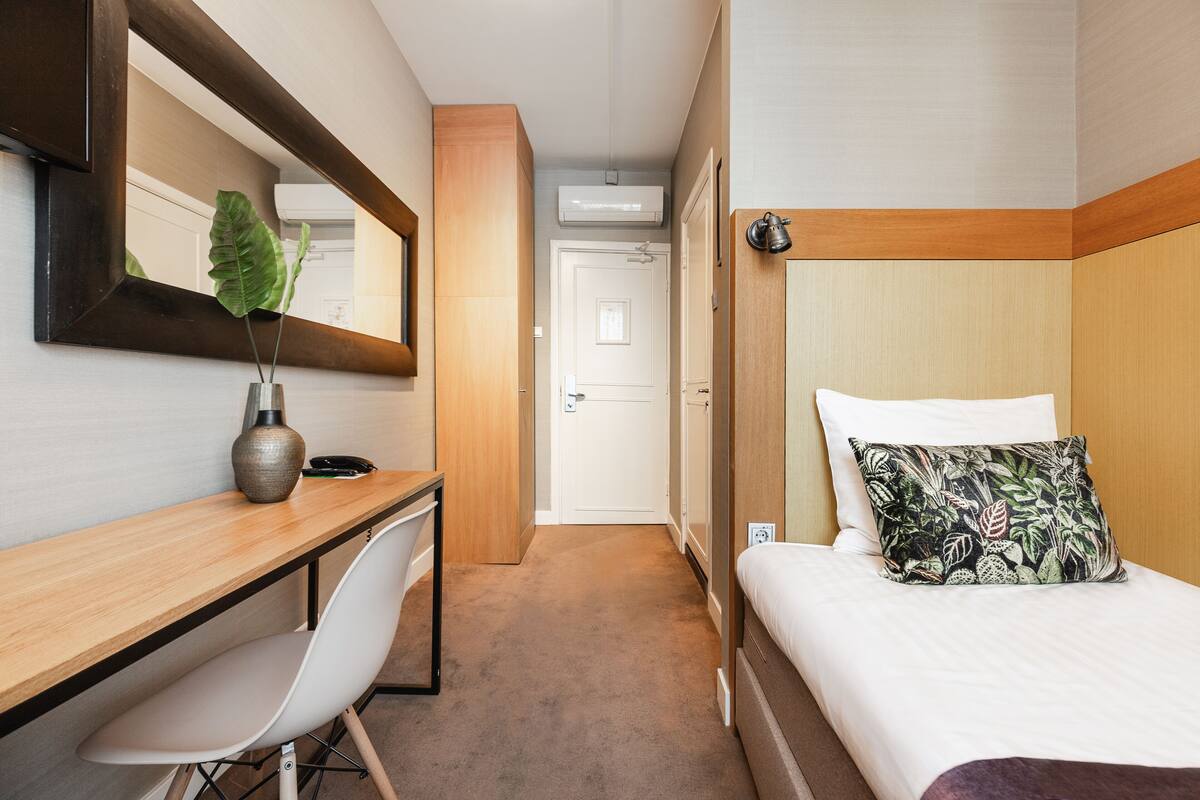 A hotel room with a wooden desk, a white chair, a bed with a patterned pillow, and a large mirror on the wall.