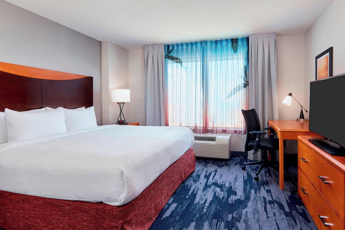 A hotel room with a large bed, a desk with a chair, a television, an air conditioning unit, and a window with curtains.