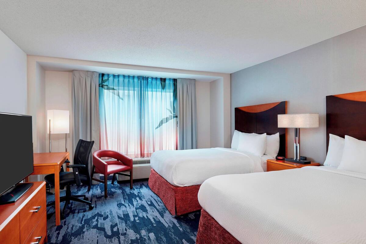 A hotel room with two beds, a desk with a chair, a TV, and a window with curtains.
