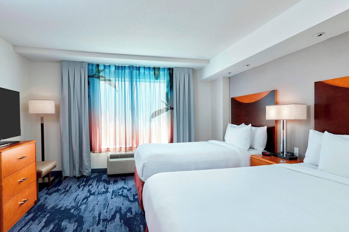 A hotel room with two beds, a desk, a chair, a TV, and a window with curtains.