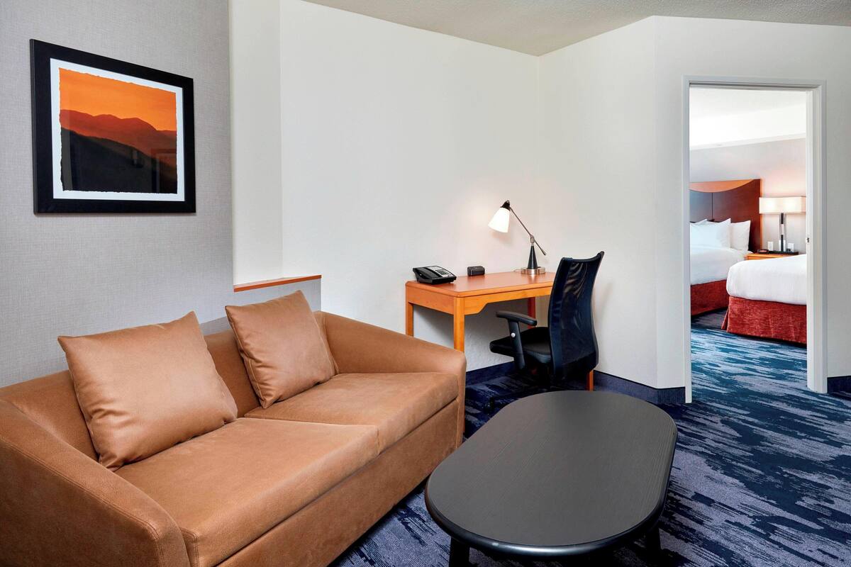 A hotel room with a brown sofa, a desk with a lamp, and a bed with red bedding.