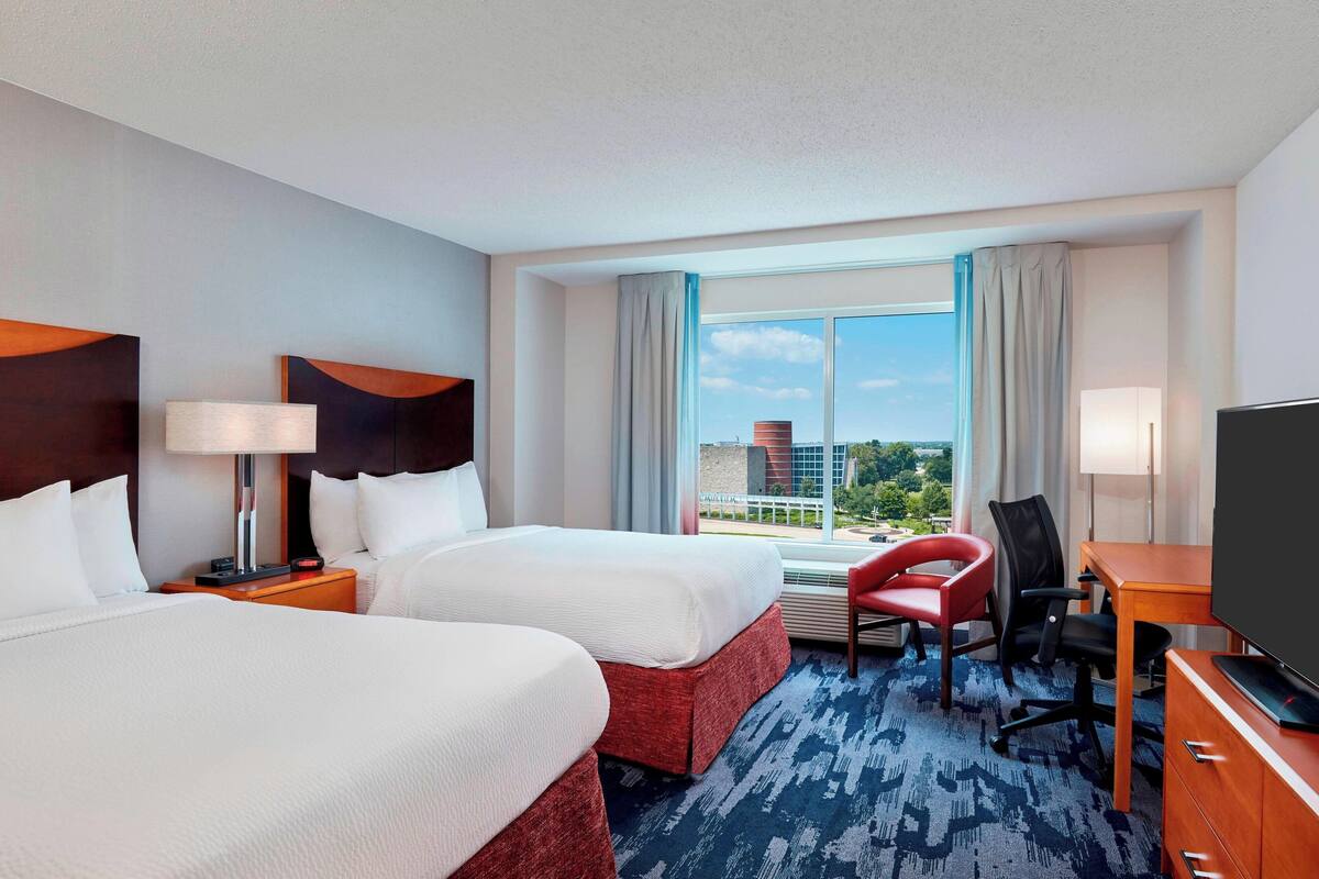 A hotel room with two beds, a desk, a TV, and a view of the city.