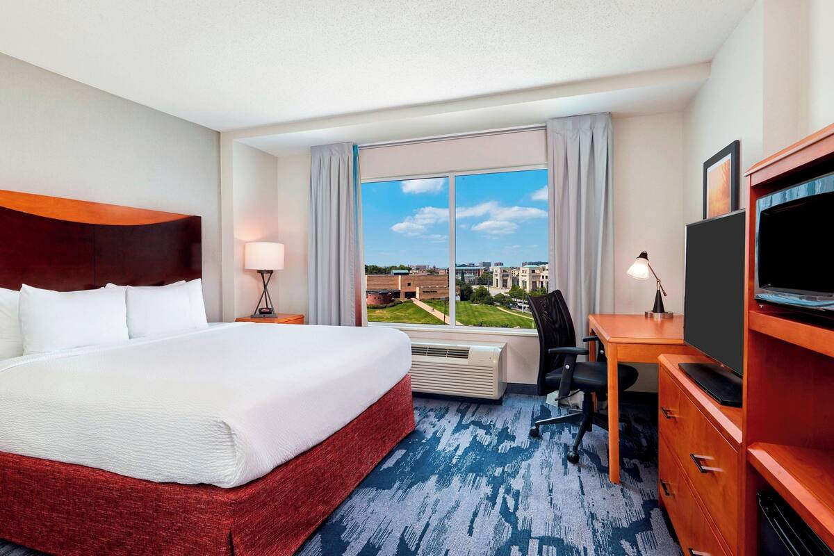 A hotel room with a large bed, a desk with a computer, a TV, and a window with a city view.