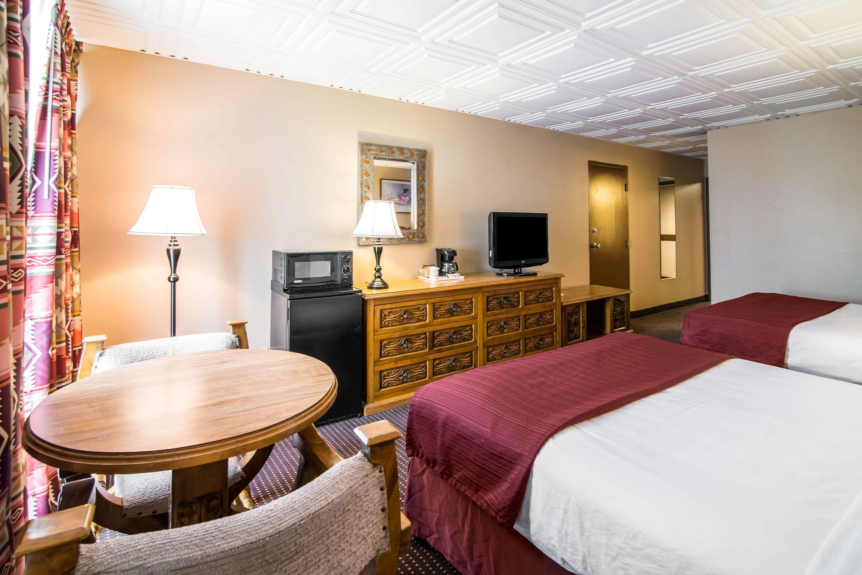 Una habitación de hotel con una mesa de madera, dos camas con ropa de cama roja y blanca, un televisor, un aparador con cajones, un espejo y una cortina estampada.