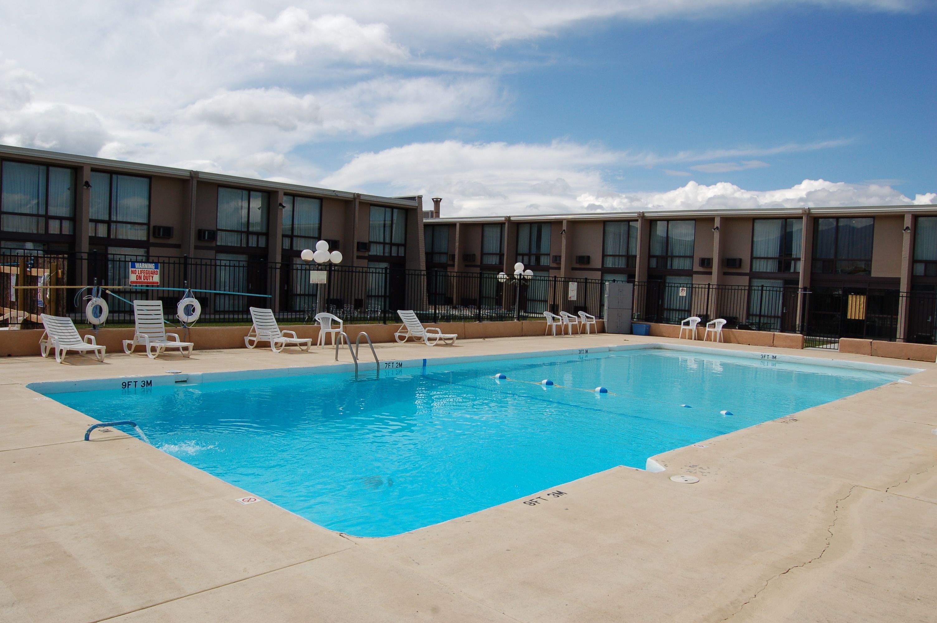 A rectangular swimming pool with a clear blue water, surrounded by a concrete deck. There are white lounge chairs aligned along the pool's edge.