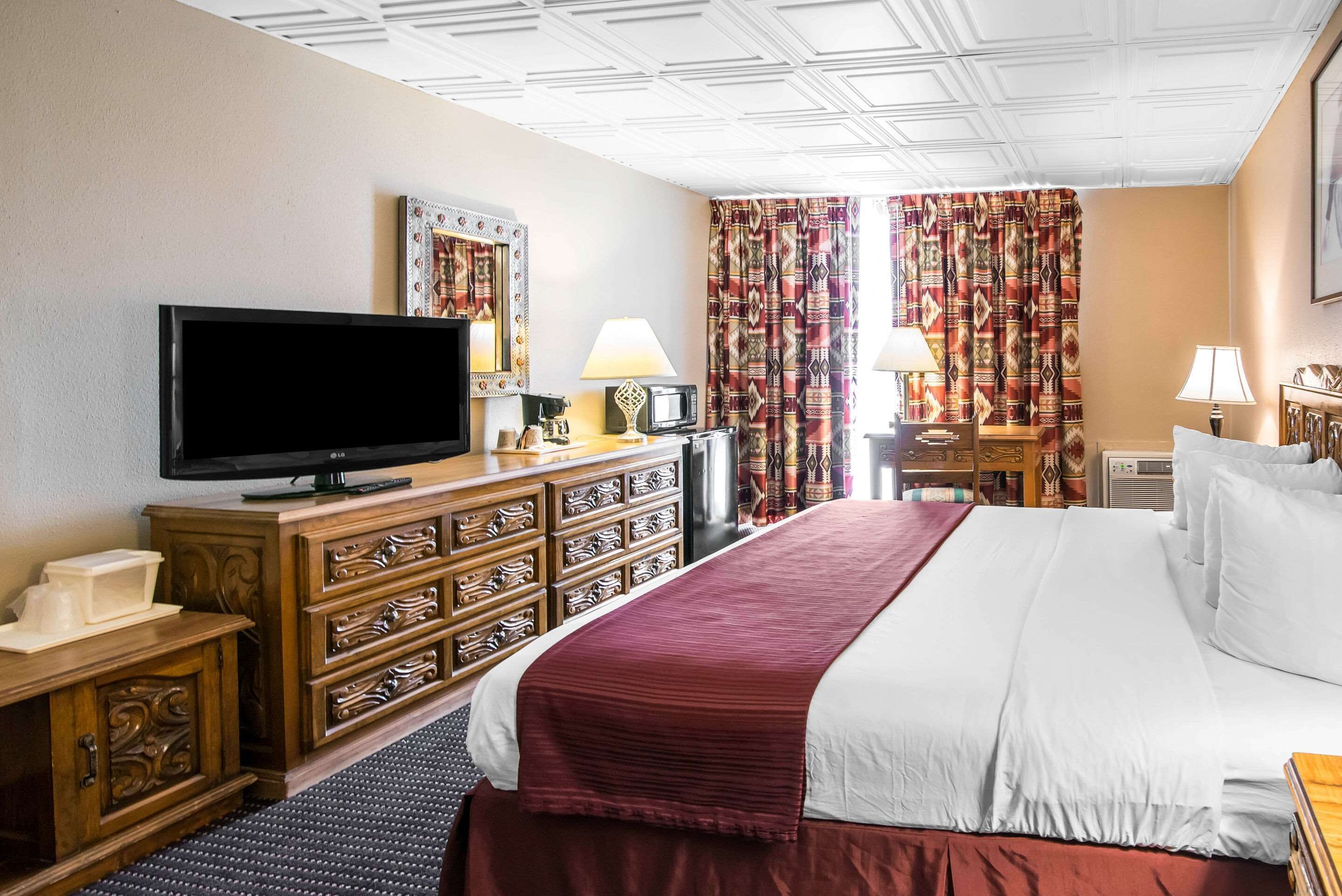 Ein Hotelzimmer mit einem großen Bett, einem Flachbildfernseher, einer Holzkommode und gemusterten Vorhängen.