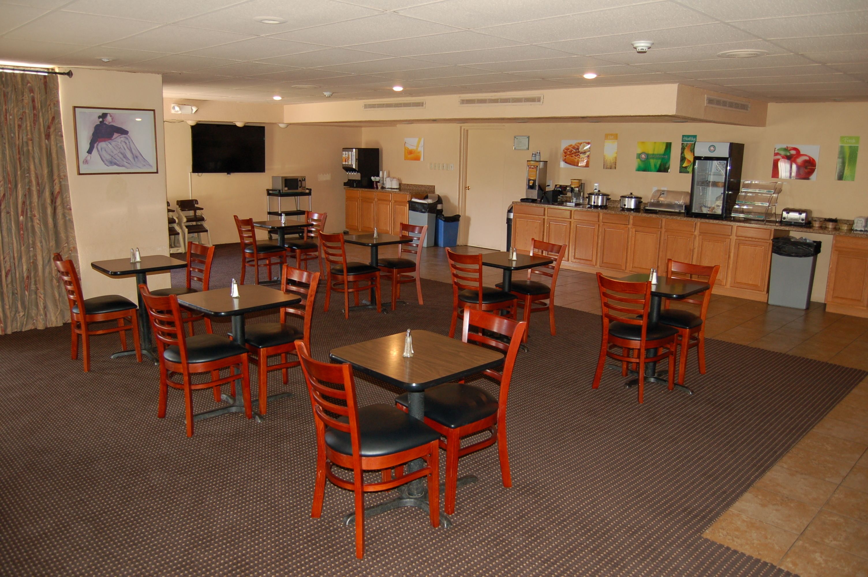 A hotel dining area with multiple tables and chairs, a kitchen area with appliances, and a television mounted on the wall.