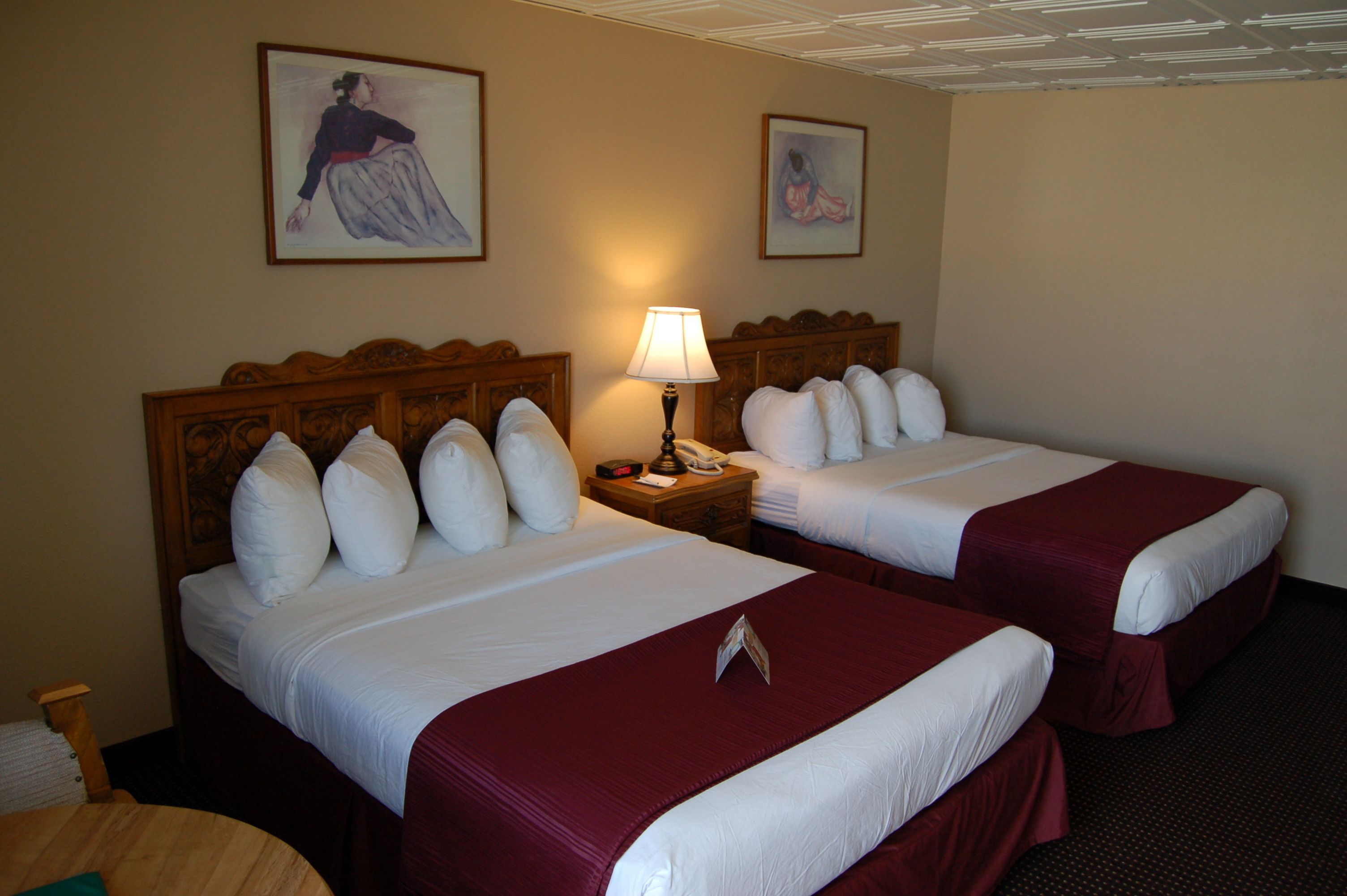 A hotel room with two beds, each with white linens and red bedspreads. There is a wooden headboard, a nightstand with a lamp, and two framed pictures on the wall.