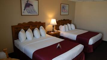 A hotel room with two beds, each with white linens and red bedspreads. There is a wooden headboard, a nightstand with a lamp, and two framed pictures on the wall.