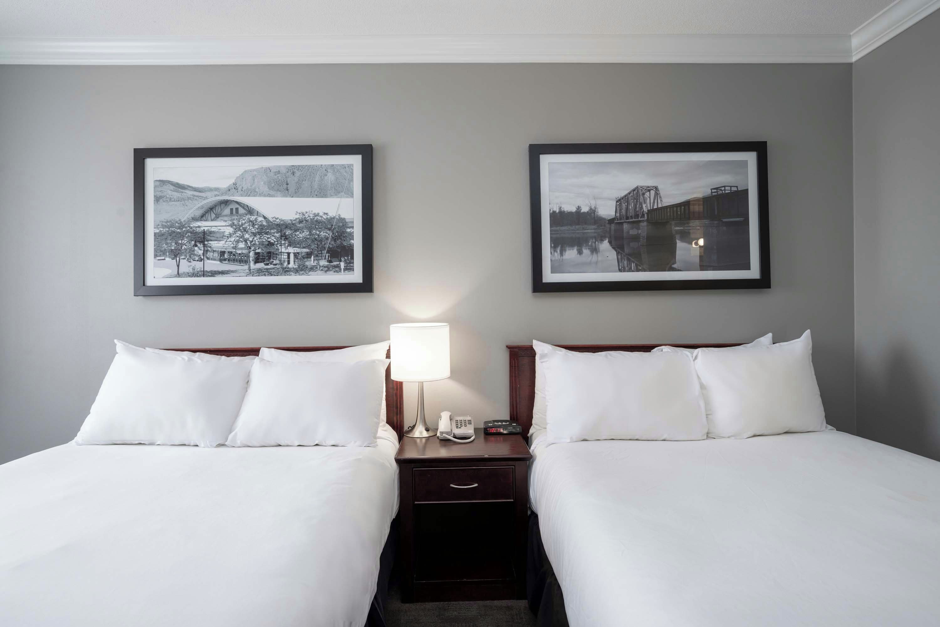 Habitación de hotel con dos camas, una mesita de noche con lámpara y dos cuadros en la pared.