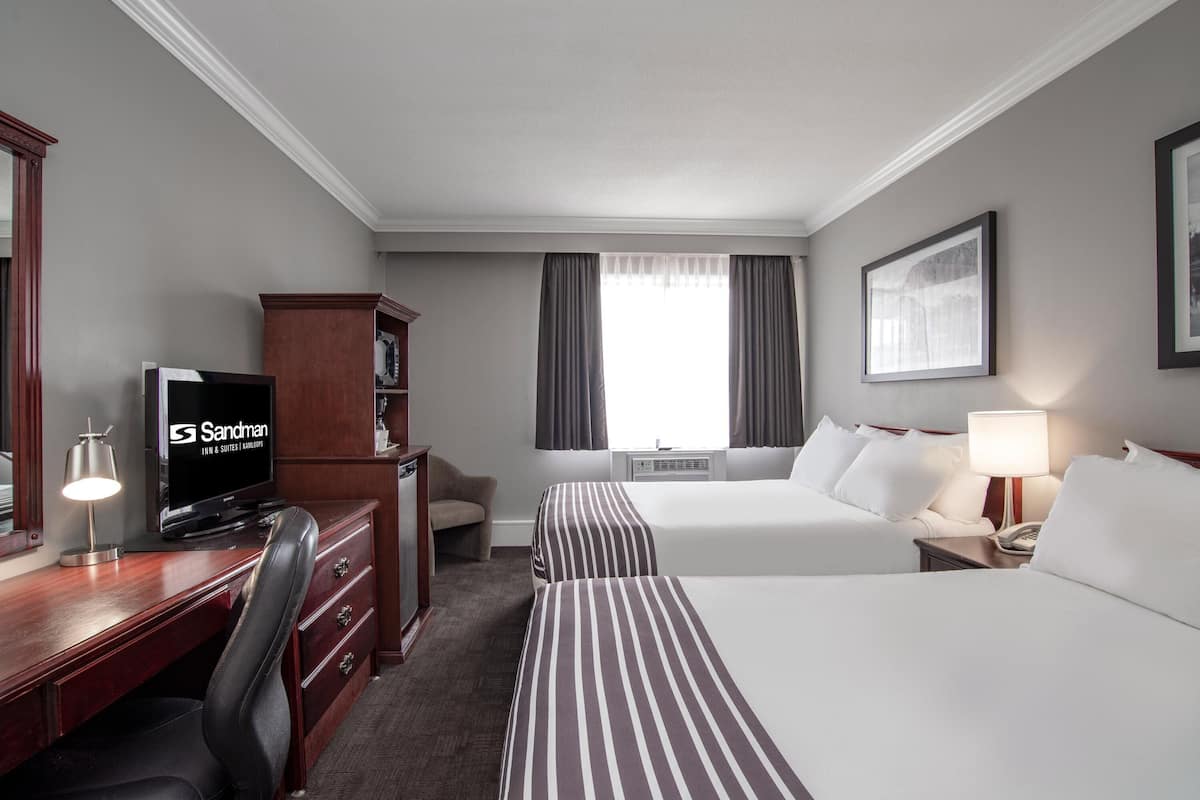 A hotel room with a desk, television, two beds, a chair, and a window with curtains.
