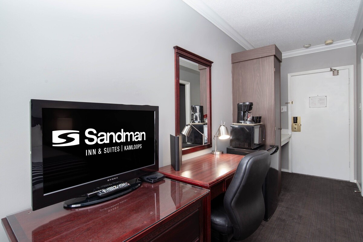 A hotel room with a TV, a desk, a chair, a mirror, and a door.
