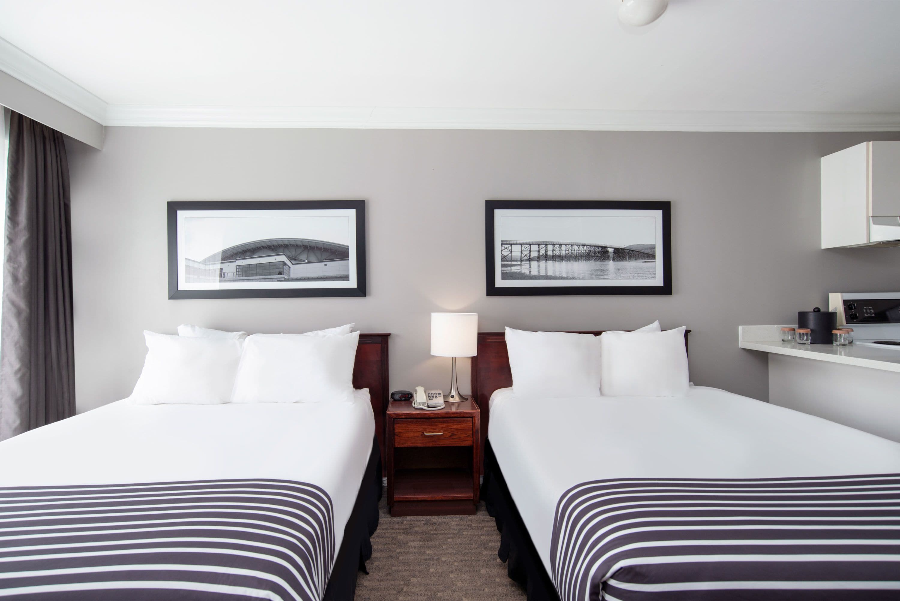 A hotel room with two beds, a nightstand with a lamp, framed pictures on the wall, and a small desk with a phone and a cup.