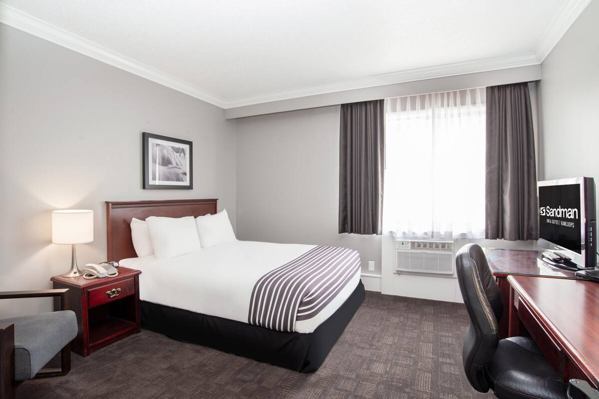 A hotel room with a large bed, a desk with a computer, a chair, a nightstand with a lamp, and a window with curtains.