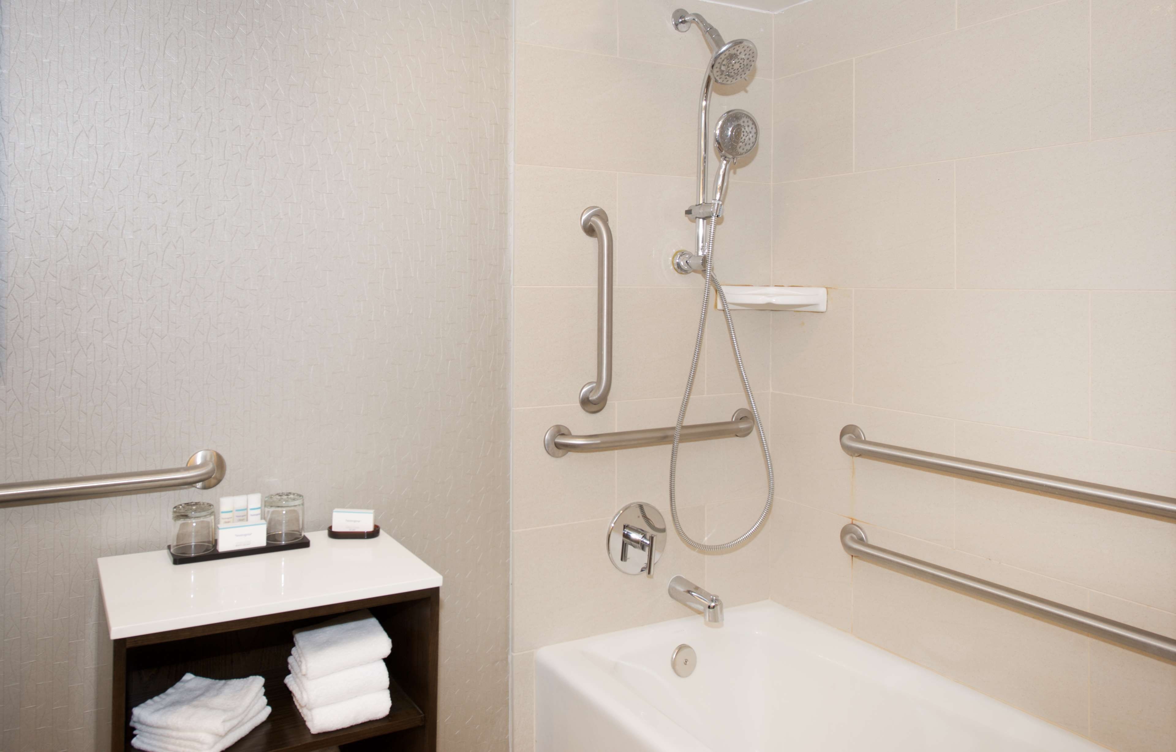 A bathroom with a bathtub, a shower with a handheld showerhead, and grab bars. There is a shelf with towels and toiletries.