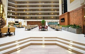 Un amplio vestíbulo de hotel con varias zonas de restauración, un área de estar central y una escalera que conduce a los pisos superiores.